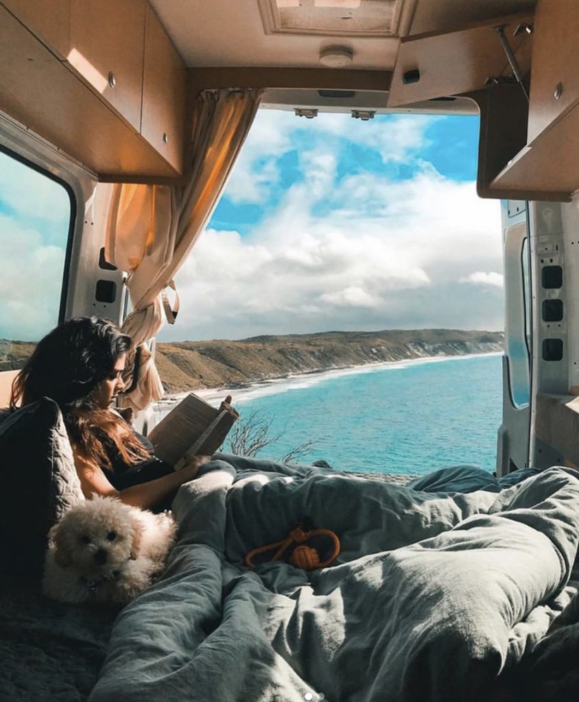 Woman sat in bed in her camper van with her dog, view of the ocean.