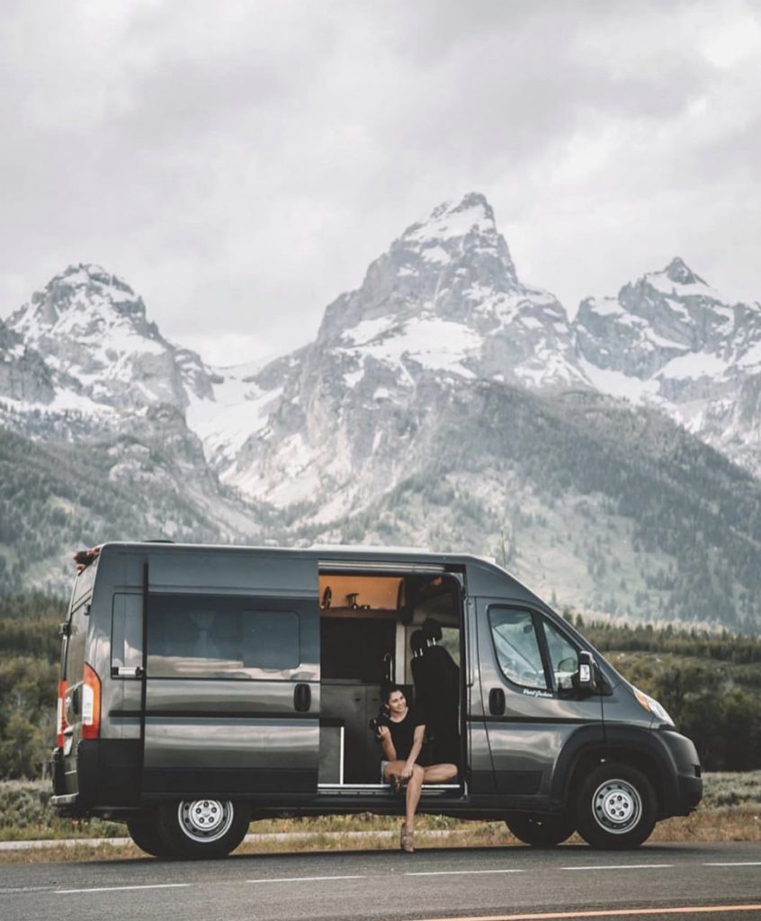 Women sat on the steps of her van in front of mountains. 