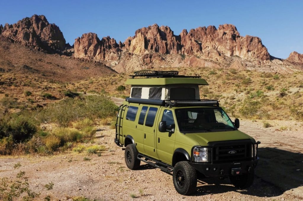 Green Sportsmobile Classic 4x4 with pop top roof open against back drop of Rocky Mountains. 
