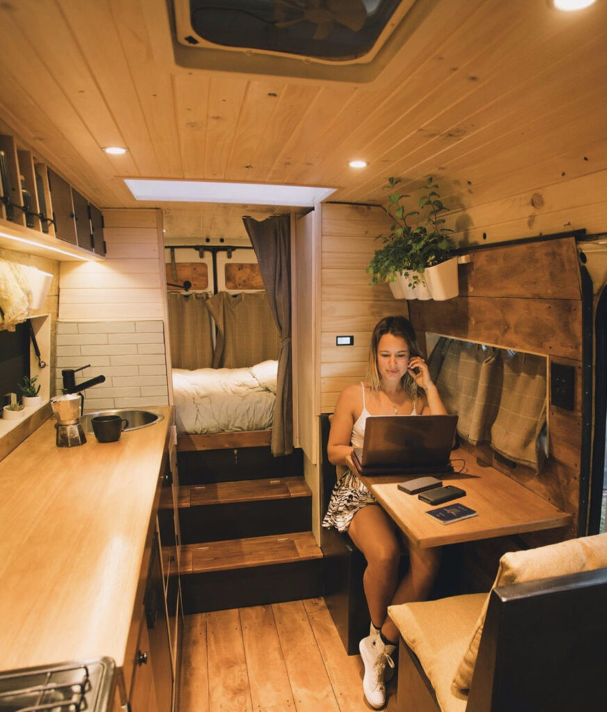 Woman sat at desk in van with bed in separate room at back and kitchen in front 